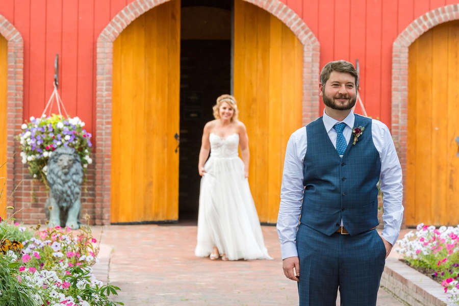 Groom in a blue plaid vest sharp in the foreground while the bride emerges from bright red barn doors with yellow arches and flower beds behind her