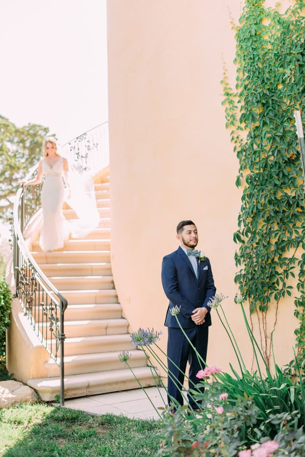 Groom in a navy suit and mint bow tie waiting at the base of a curved stone staircase as the bride descends above him, against an ivy-covered terracotta wall
