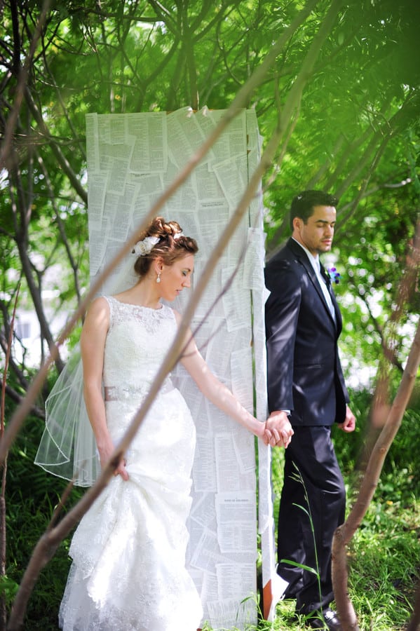 Bride and groom holding hands on opposite sides of a tall DIY door covered in Bible pages, eyes closed, surrounded by greenery