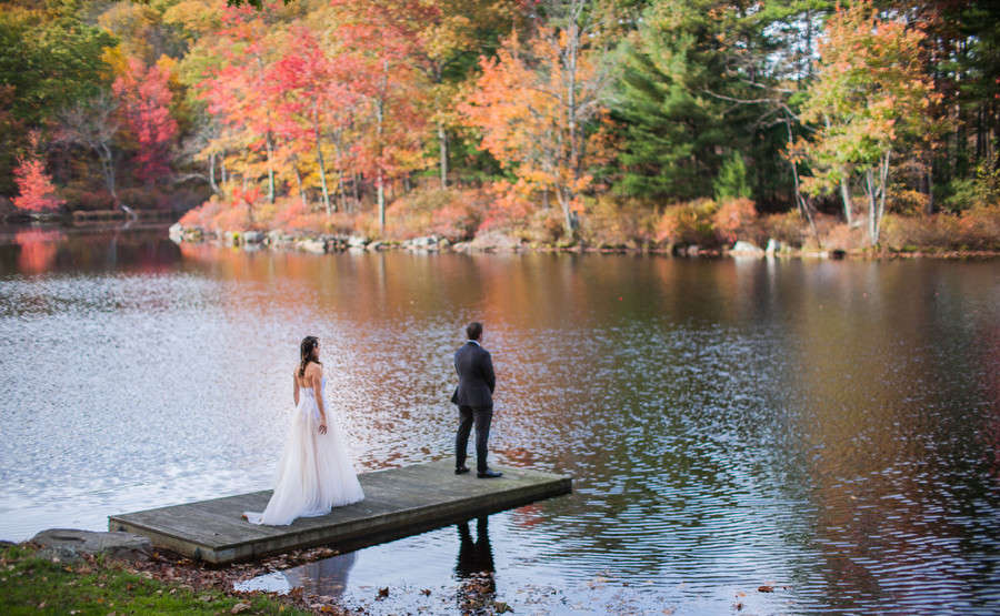 Wide first look of the groom standing at the end of a lake dock as the bride approaches, vivid autumn foliage reflected in the still water