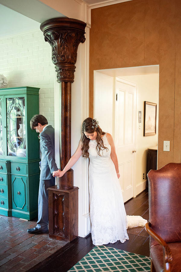 Bride and groom on opposite sides of a carved wooden column holding hands and bowing their heads, in a vintage home interior