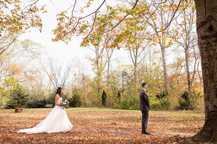 Wide first look of the bride and groom facing each other across a clearing covered in fall leaves, golden autumn canopy overhead