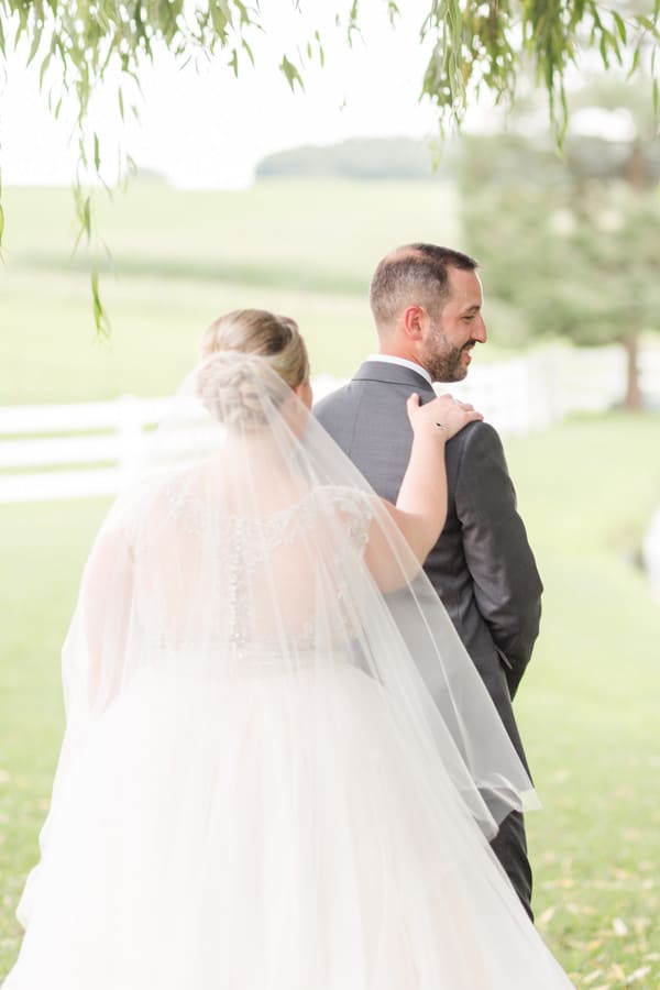 Groom in a grey suit turning and smiling during the first look, seen over the bride's veil and framed by draping willow branches at a farm