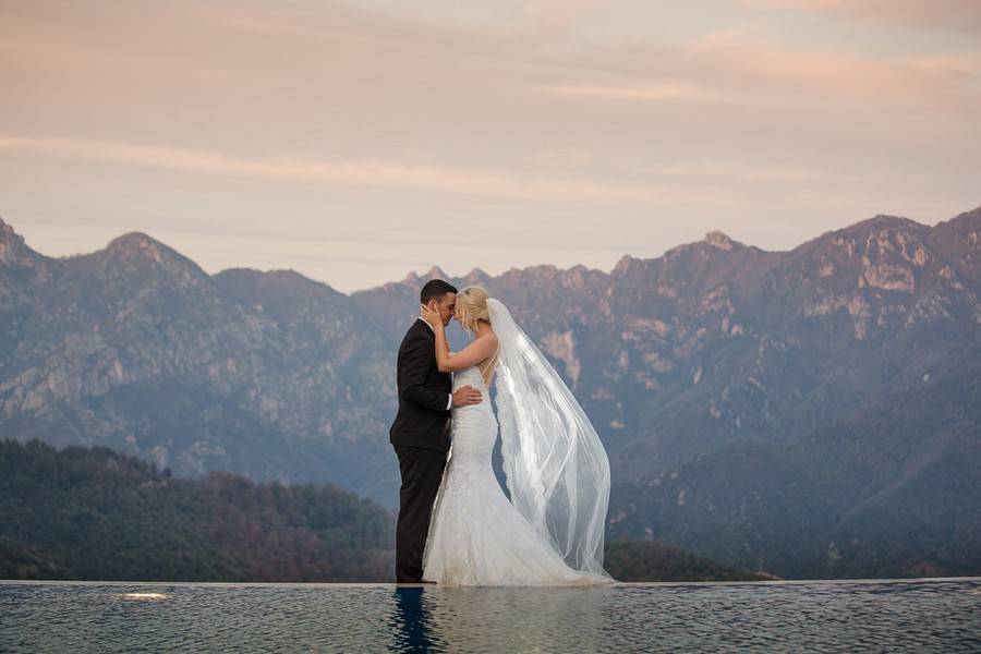 Couple with foreheads touching at an infinity-pool edge, the long veil billowing against mountains at dusk