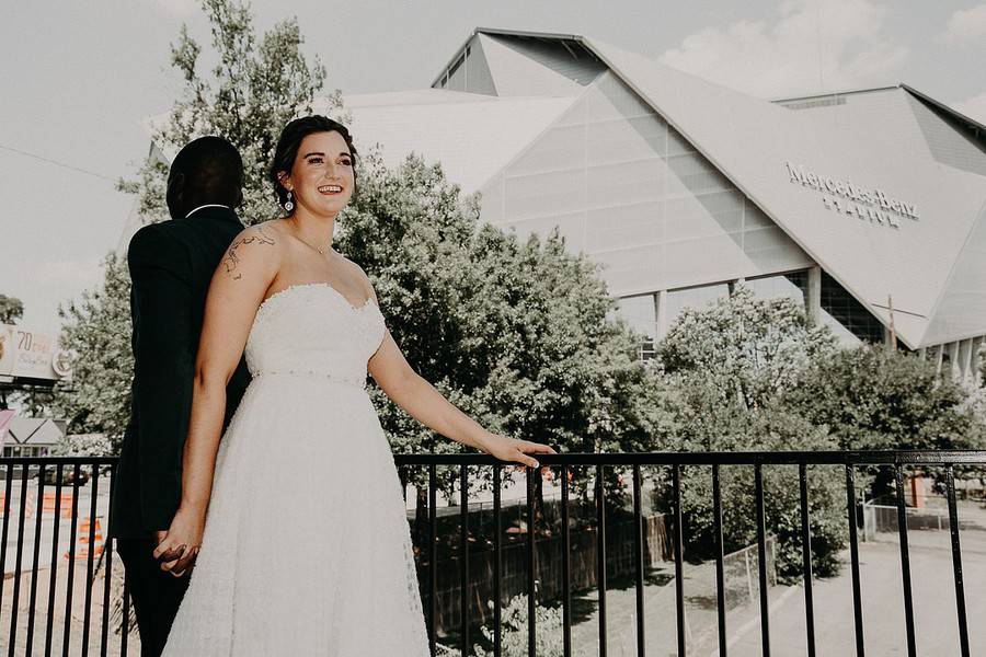 Bride and groom standing back-to-back holding hands on a railing, smiling, with Mercedes-Benz Stadium in the Atlanta skyline behind them