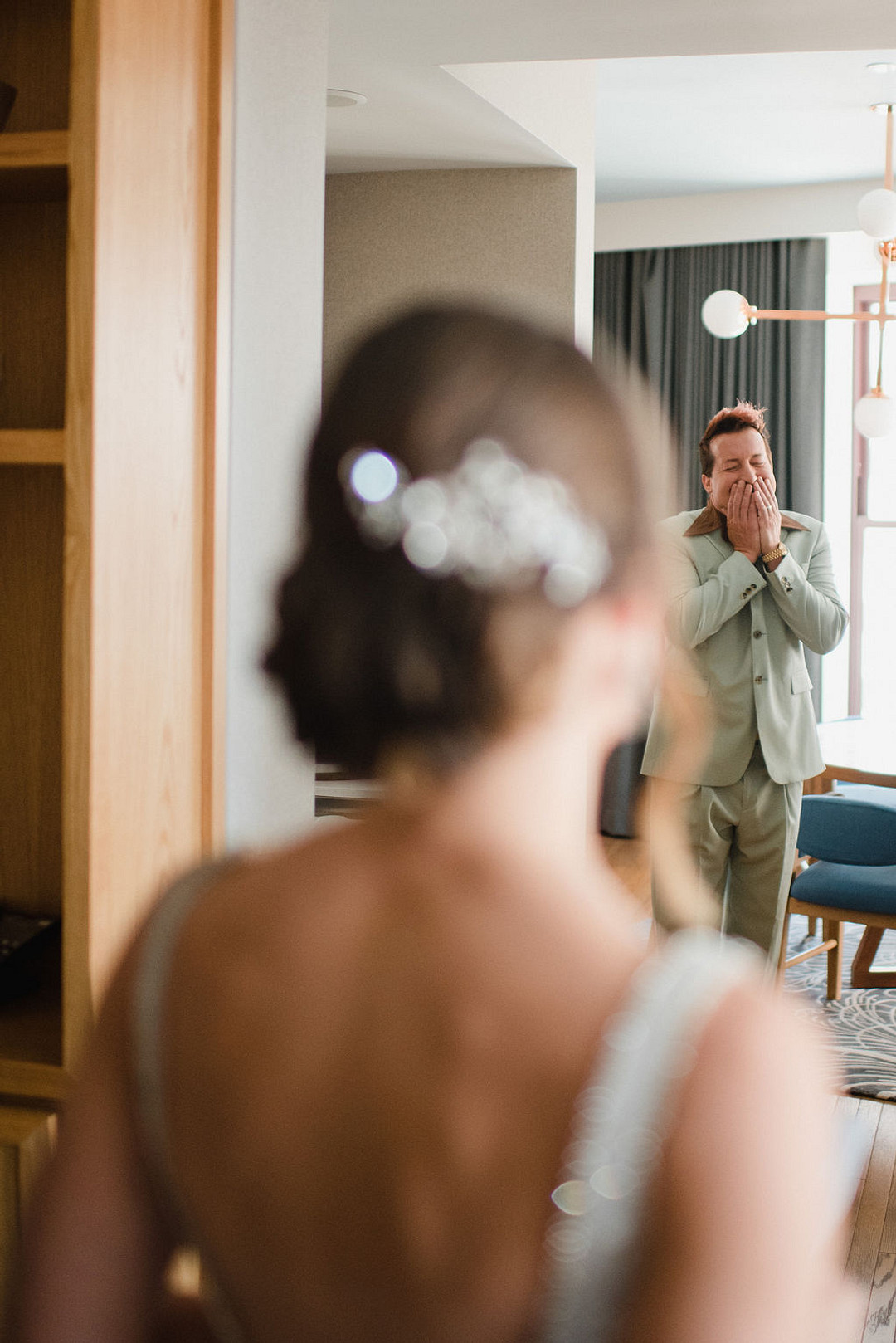 Groom in a sage green suit covering his mouth with both hands as he reacts to the bride during their first look, back of the bride's jeweled updo soft in the foreground
