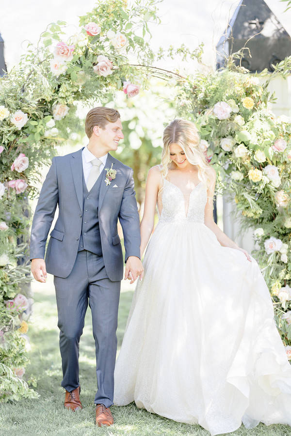 Bride and groom walking hand in hand through a lush pastel floral arch on grass
