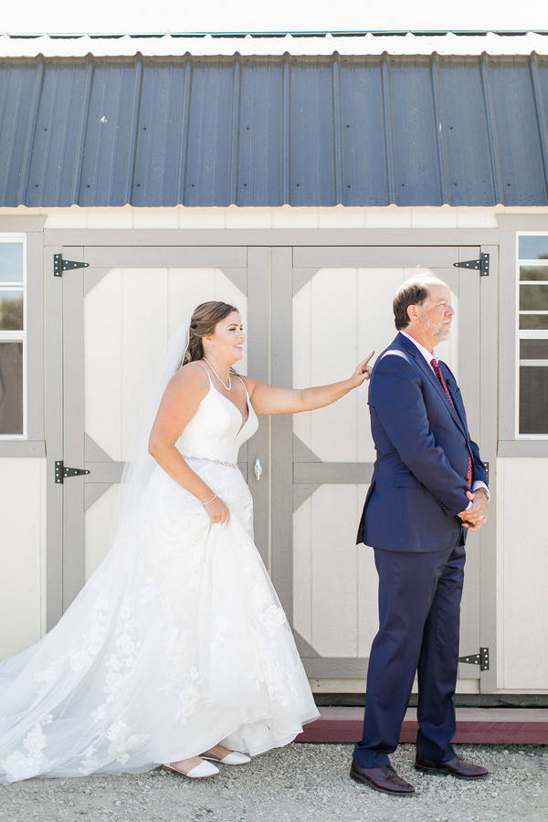 Bride in a lace ballgown and cathedral veil tapping her father's shoulder for a first look in front of grey barn doors