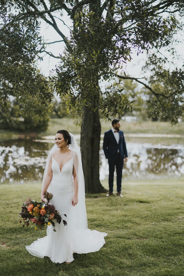 Bride sharp in the foreground holding a burnt-orange autumn bouquet while the groom waits under a tree by a pond in the soft background