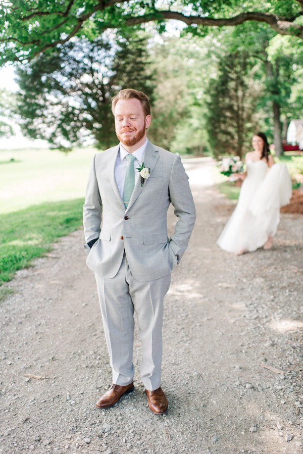 Groom in a light grey suit and sage green tie waiting with his eyes closed on a tree-lined gravel farm lane as the bride approaches behind him