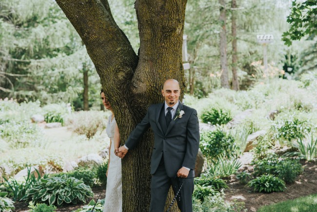 Bride and groom holding hands on opposite sides of a large tree trunk for a first touch, neither one looking, in a lush green garden