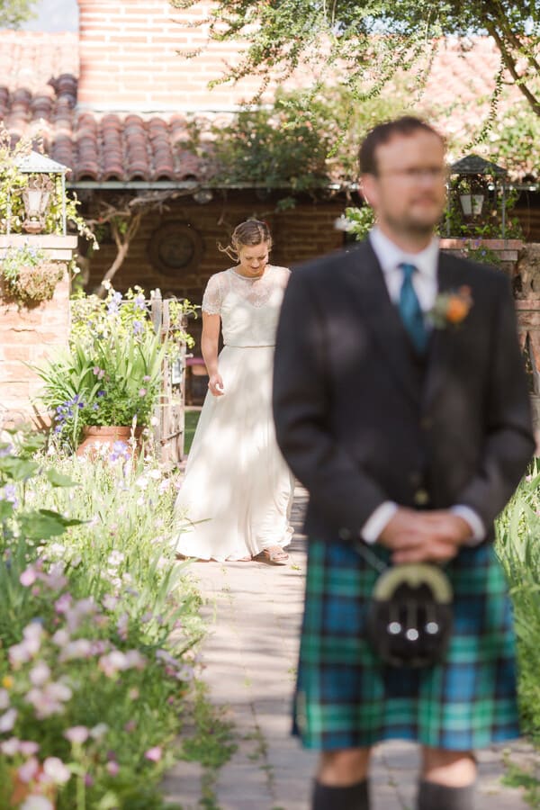 Groom's green tartan kilt and sporran filling the soft foreground while the bride is in sharp focus approaching down a wildflower garden path past an adobe building