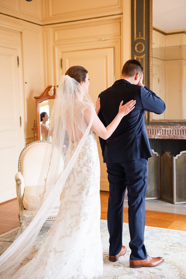 Bride with a long cathedral veil and beaded gown facing the groom in a navy suit who wipes his eye during an indoor first look, her reflection caught in an ornate mantel mirror