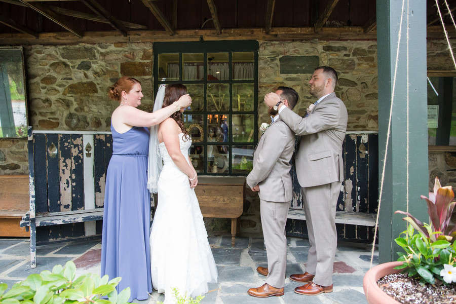 A bridesmaid in a periwinkle dress and a groomsman in a tan suit each covering the bride's and groom's eyes, walking them together for a first look in front of a stone barn wall with a LOVE sign