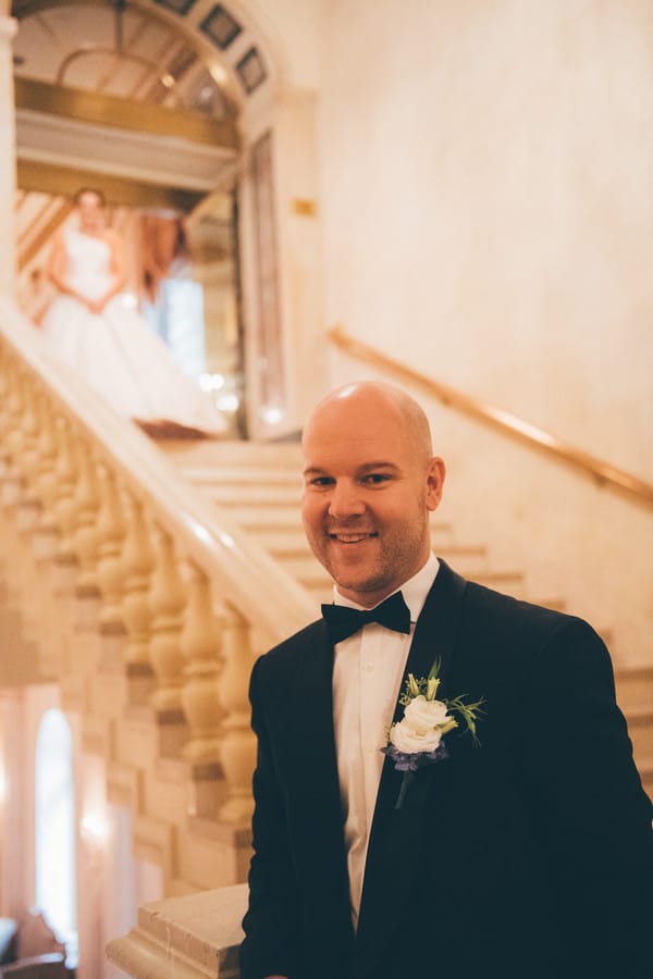 Groom in a navy suit waiting at the bottom of a grand stone staircase as the bride descends, classic hotel interior