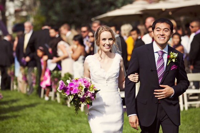 Bride and groom walking arm in arm down a grassy recessional aisle past seated guests
