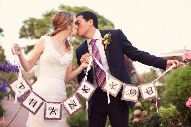 Couple kissing while holding the ends of a hanging THANK YOU banner in a garden