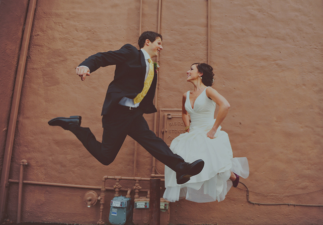 Bride and groom jumping mid-air facing each other against a flat orange-painted wall