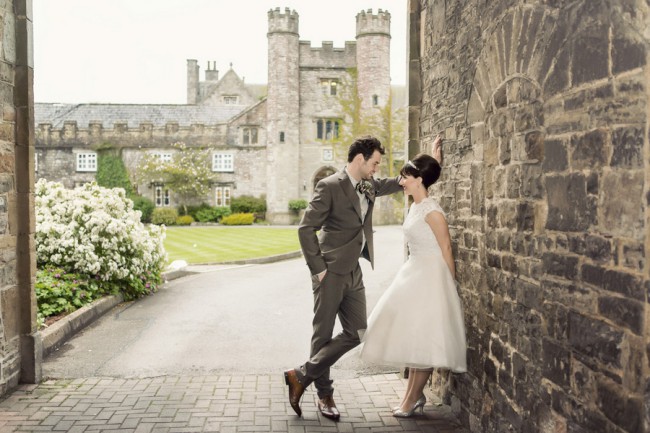 Couple leaning against a stone wall, gazing at each other with a manor house behind them