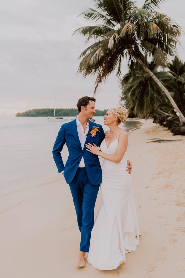 Bride and groom strolling arm around each other along a tropical beach under a palm tree