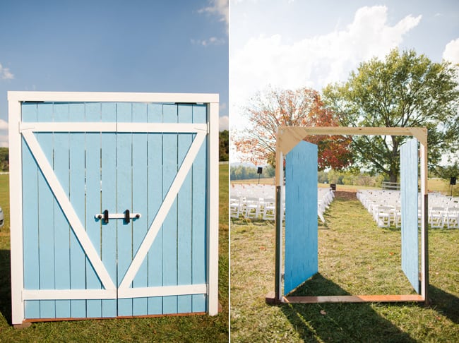 Freestanding bright blue barn door gateway opening onto a chair-lined ceremony aisle with a fall tree behind