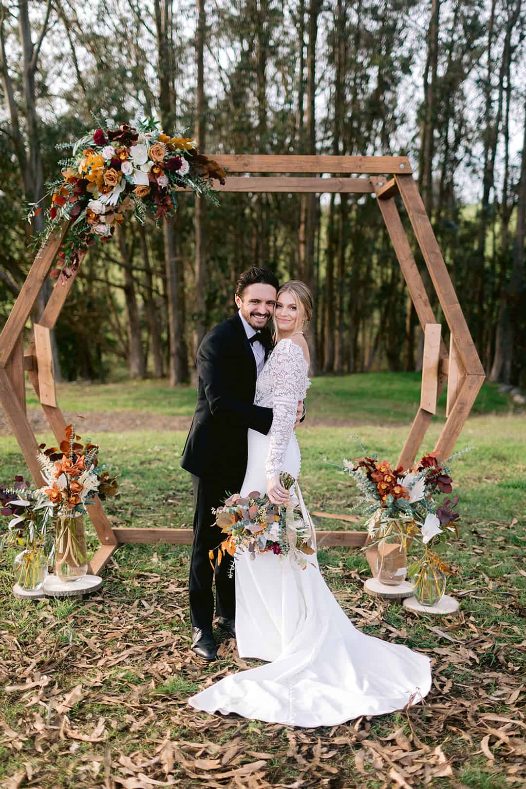 Hexagonal wooden wedding arch with rust and burgundy autumn florals on the top corner, couple standing in front in a eucalyptus grove