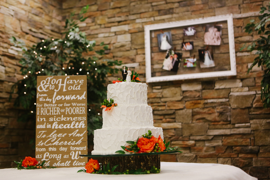 Three-tier white textured buttercream cake with orange roses and green berries on a wood slab against a stone wall