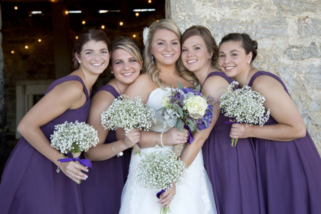 Bridesmaids in plum purple gowns holding baby's breath bouquets in a string-lit barn