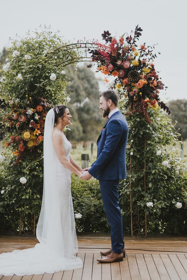 Iron wedding arbor covered in greenery white roses and burnt-orange native flowers, couple holding hands on a wood deck