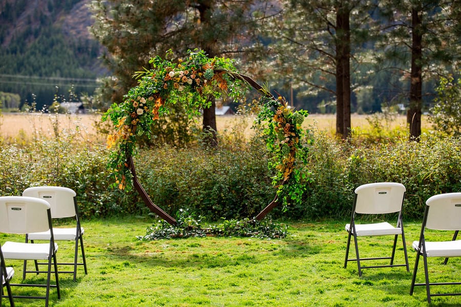 Round dark wood wedding arbor draped in greenery and marigold blooms in a meadow with mountains behind