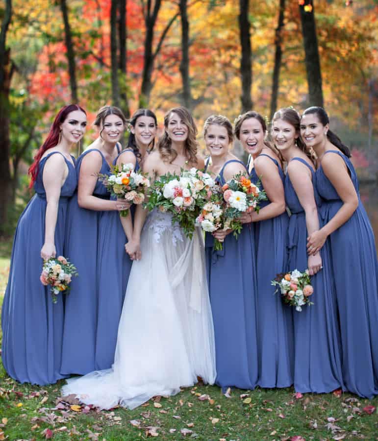 Eight bridesmaids in slate blue gowns in front of a bright autumn forest