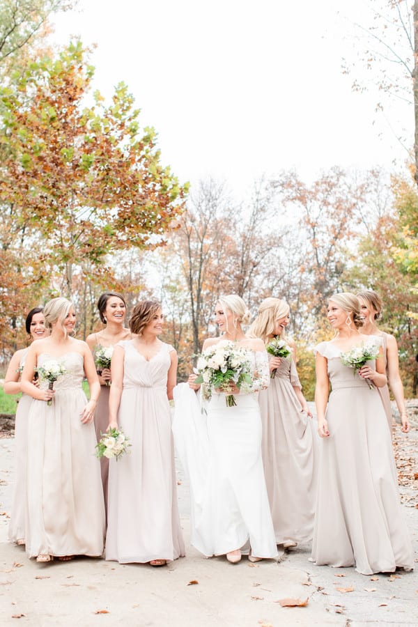 Bridesmaids in taupe and champagne floor-length gowns walking on a leaf-covered path