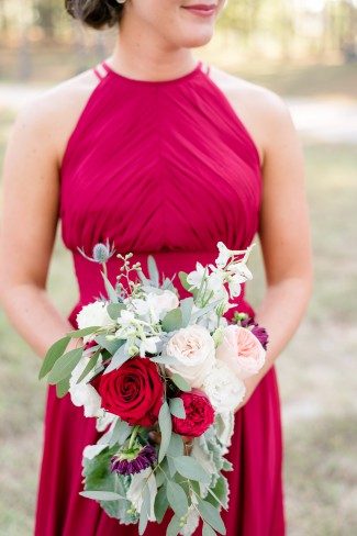 Close-up of a burgundy halter bridesmaid dress with a red rose and eucalyptus bouquet