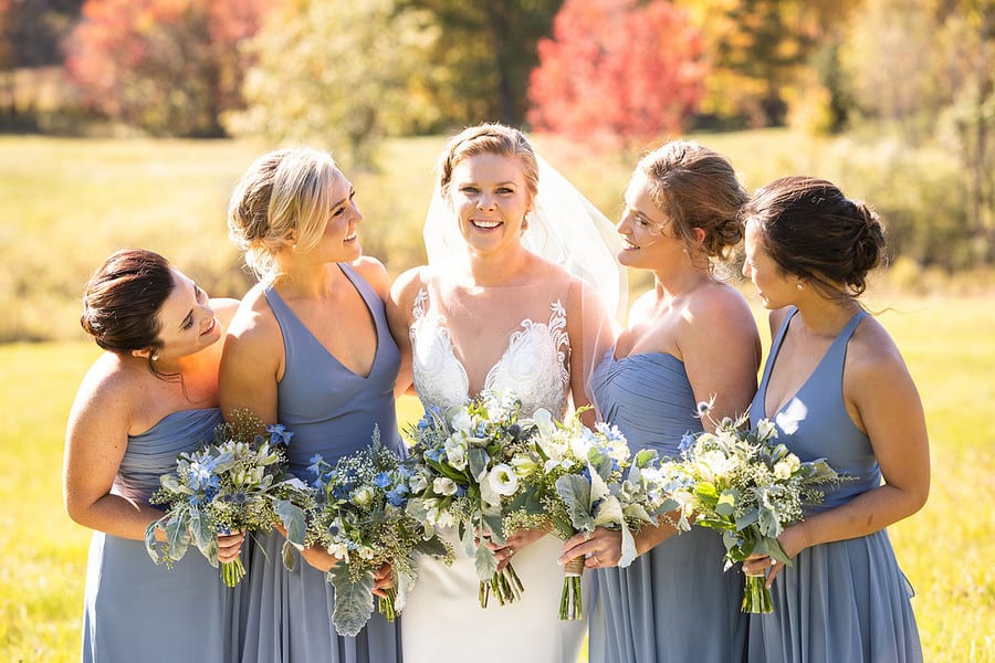 Bridesmaids in dusty blue gowns holding blue and white bouquets with fall trees behind