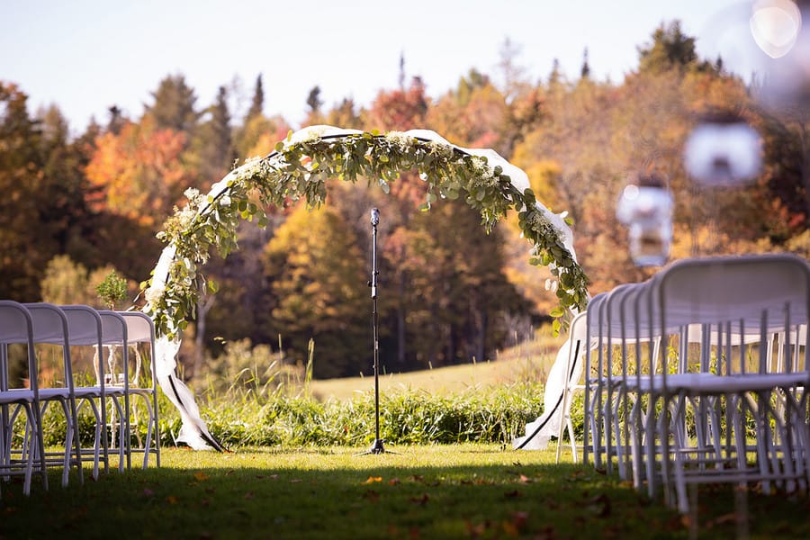 Circular wedding arch with white drape and greenery against a backdrop of orange and red fall foliage