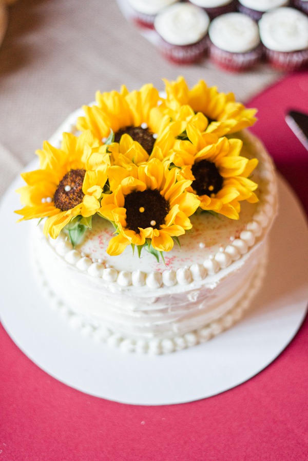 Single-tier white buttercream cake topped with fresh yellow sunflowers, red velvet cupcakes behind