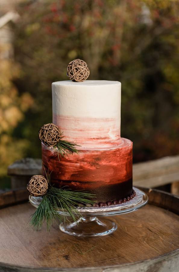 Two-tier wedding cake with a white-to-burgundy ombre finish, twig balls, and pine sprigs on a wood barrel