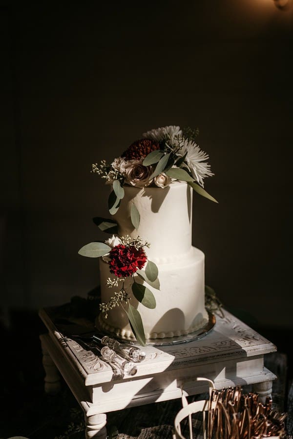 Two-tier white wedding cake with deep burgundy dahlias, mauve roses, and eucalyptus in moody light