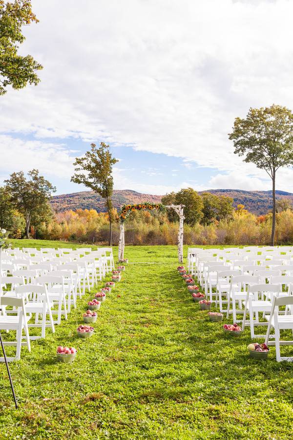 White birch log wedding arch with a sunflower garland at the end of a grass aisle lined with apples and fall foliage mountains behind