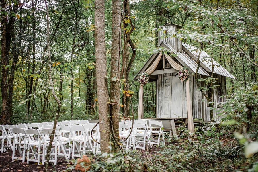 Weathered wood chapel with a fabric-draped doorway and blush florals set in the forest with white chairs