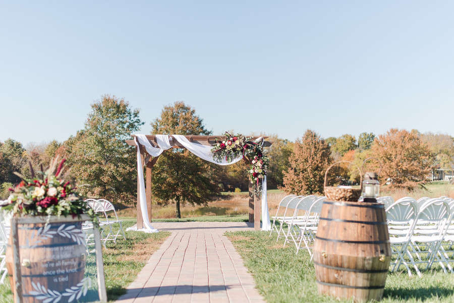 Square timber wedding arch with white draped fabric and a burgundy floral cluster at the end of a brick aisle with wine barrels