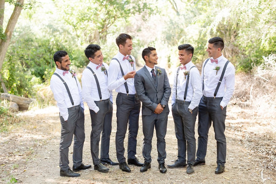 Groom in a grey three-piece suit surrounded by groomsmen in white shirts grey suspenders and blush bow ties