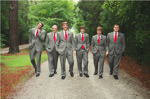 Six groomsmen in grey suits with bright red neckties walking down a gravel lane through pine woods