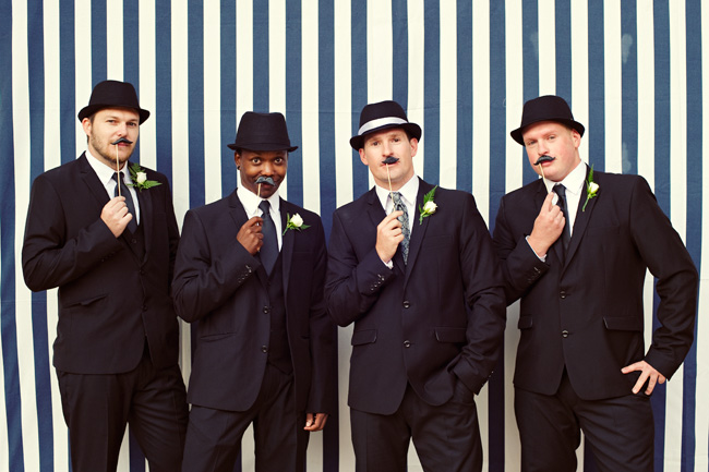 Four groomsmen in black suits wearing bowler and trilby hats holding mustache props against a striped wall