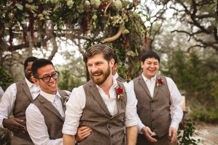 Groom and groomsmen laughing in brown herringbone waistcoats over open collar shirts with deep red rose boutonnieres