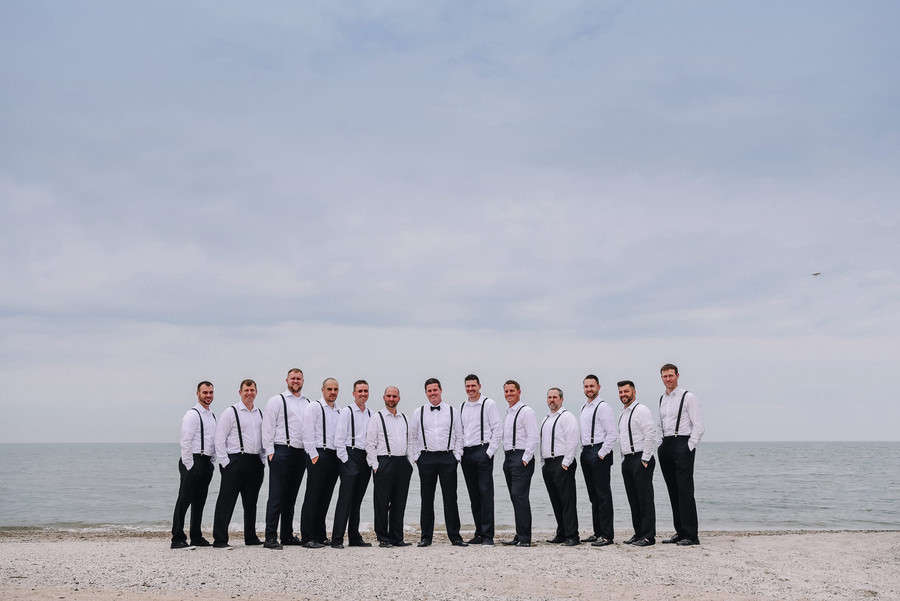 Fourteen groomsmen in white shirts black suspenders and bow ties standing in a long line on a lake beach