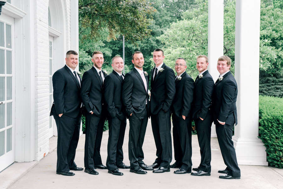 Eight groomsmen in black suits standing in a wedge formation under a white colonnade with the groom in a blush tie