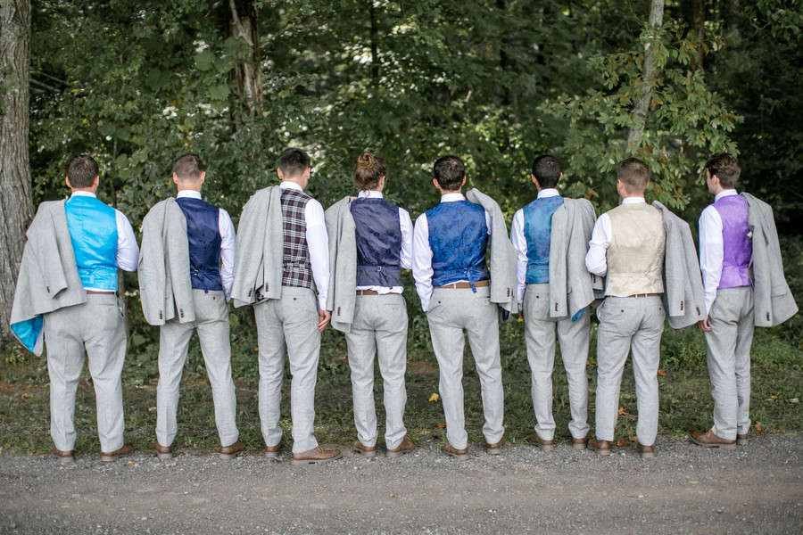 Eight groomsmen photographed from behind in grey trousers with jackets over their shoulders showing different patterned waistcoat backs