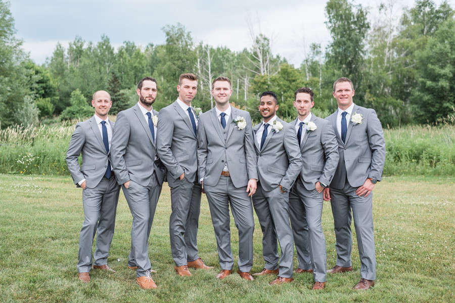 Seven groomsmen in light grey suits with navy ties and tan leather shoes standing in a summer meadow