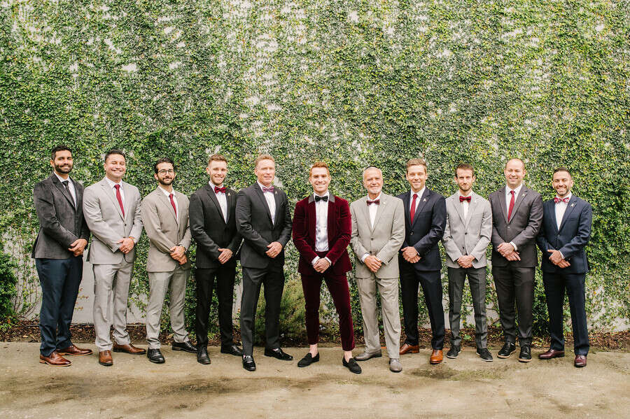 Eleven groomsmen in mismatched grey and navy suits with burgundy ties beside a groom in a burgundy velvet suit